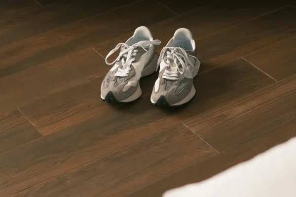 Pair of gray and white sneakers with laces on dark wood flooring beneath a white bed frame.