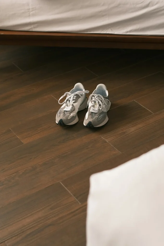 Pair of gray and white sneakers with laces on dark wood flooring beneath a white bed frame.