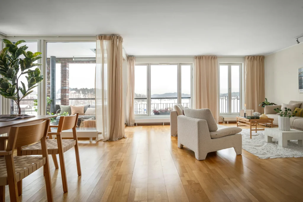 Modern penthouse living room with floor-to-ceiling windows, wooden floors, beige curtains, mid-century furniture, and waterfront views.
