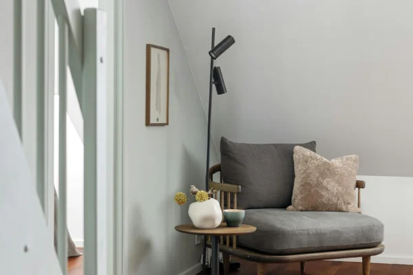 Minimalist attic room with gray armchair, wooden side table, black floor lamp, and white walls illuminated by skylight window.