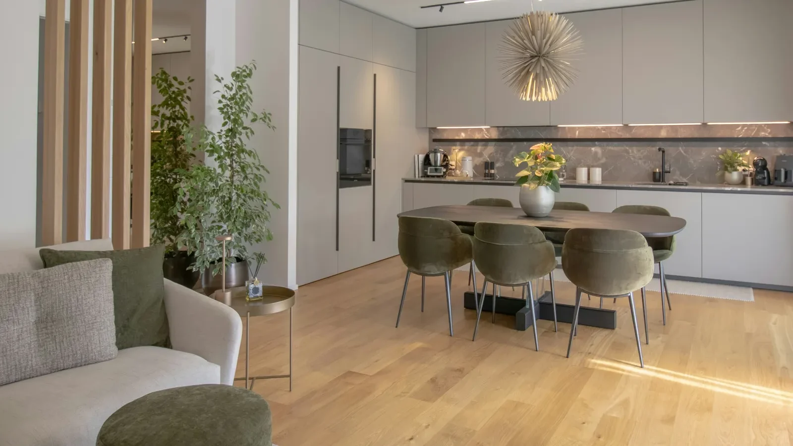 Modern open-plan apartment with kitchen, dining area, and living room featuring white cabinetry, wooden flooring, and green upholstered furniture.
