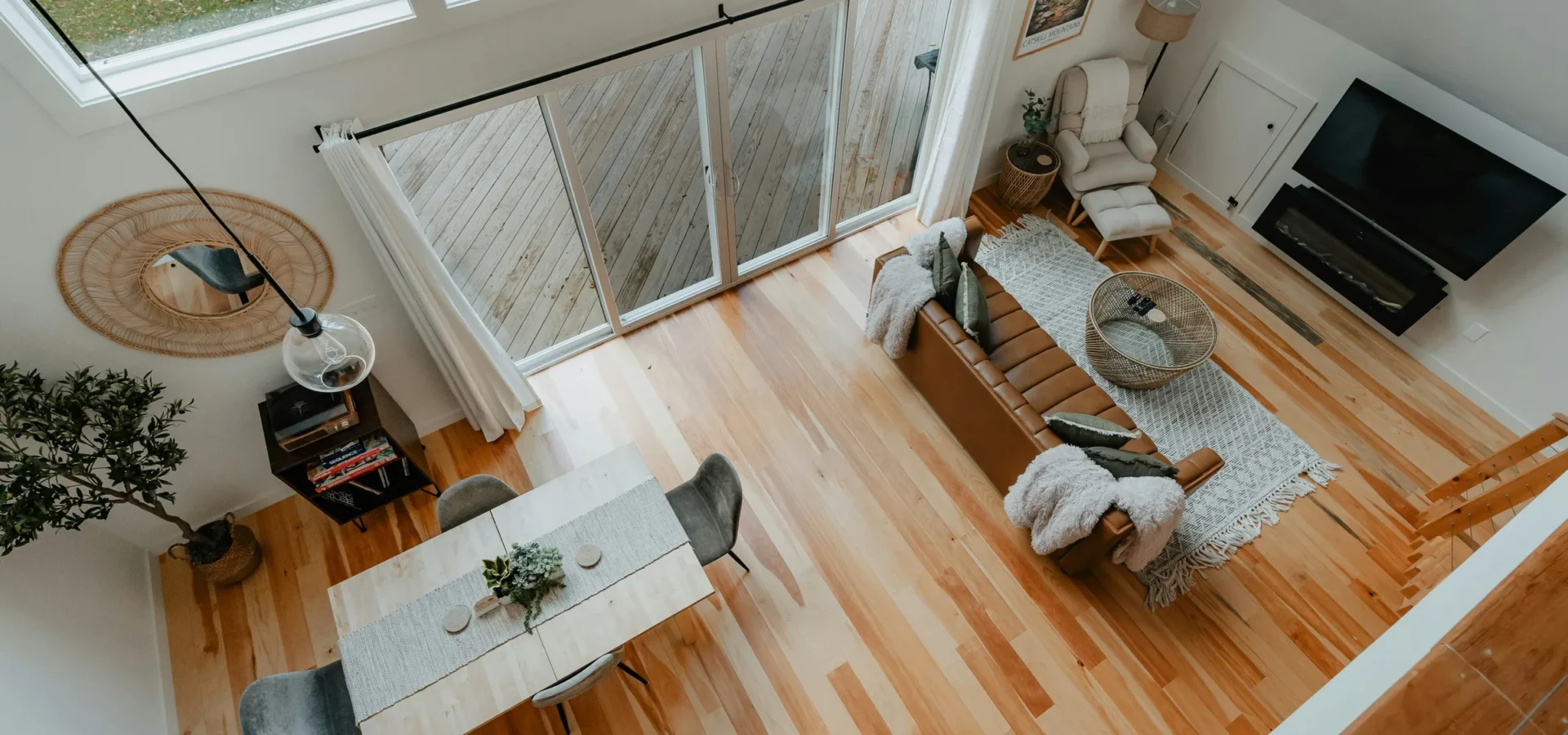 Overhead view of a modern loft apartment with wooden floors, dining table, sofa, chair, TV, and large windows overlooking greenery.