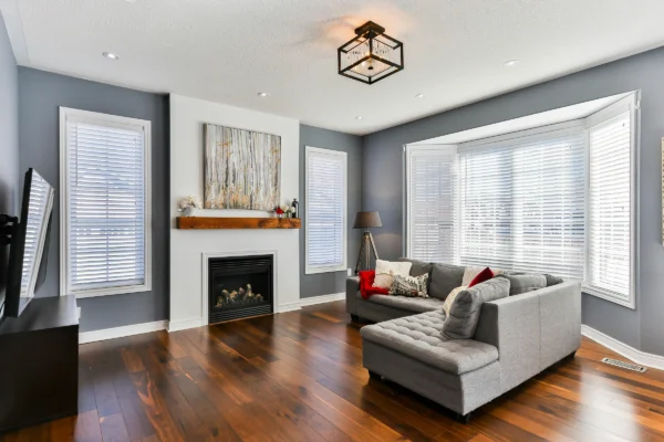 Modern living room with gray walls, white shutters, gray sectional sofa, fireplace, wood flooring, and decorative lighting.