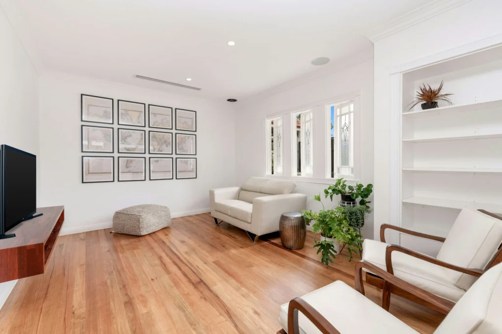 Modern living room with cream furniture, framed artwork, wooden floors, white built-in shelving, and large windows.