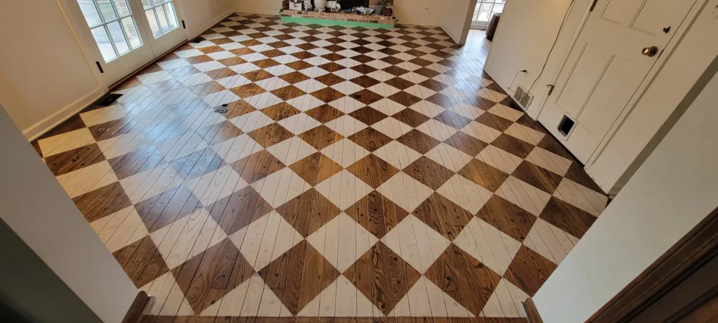 Spacious room with diamond-patterned hardwood flooring in alternating brown and light wood tones, cream-colored walls, and natural light from windows.